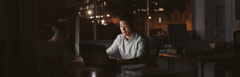 Young Asian businessman sitting at his desk working on a laptop in a dark office at night with city lights in the background