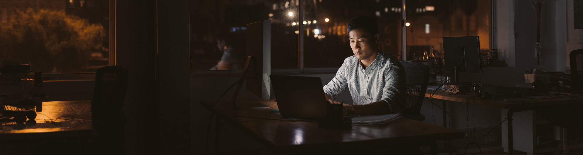 Young Asian businessman sitting at his desk working on a laptop in a dark office at night with city lights in the background