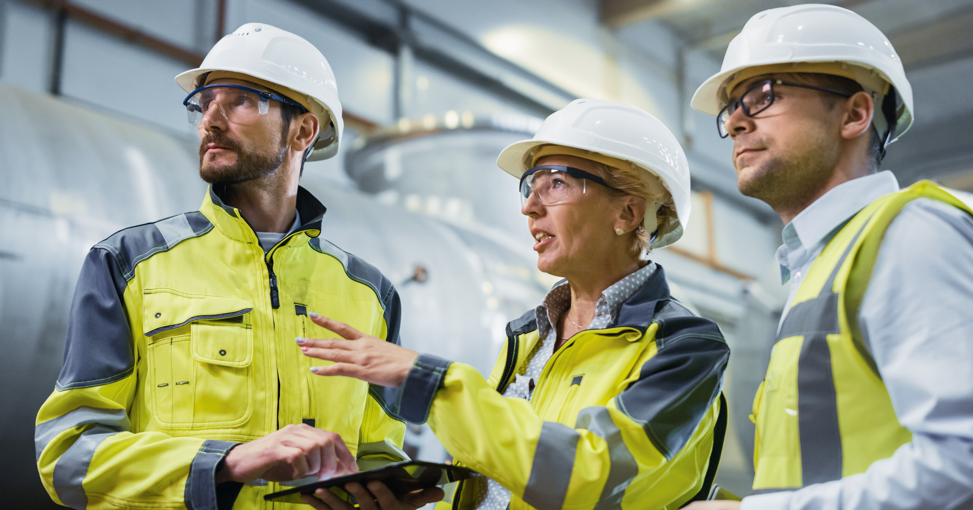 Three Heavy Industry Engineers Stand in Pipe Manufacturing Factory, Use Digital Tablet Computer, Have Discussion. Large Pipe Assembled. Design and Construction of Oil, Gas and Fuels Transport Pipeline