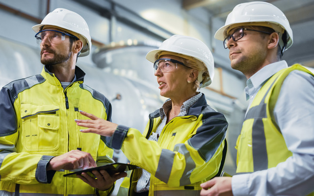 Three Heavy Industry Engineers Stand in Pipe Manufacturing Factory, Use Digital Tablet Computer, Have Discussion. Large Pipe Assembled. Design and Construction of Oil, Gas and Fuels Transport Pipeline