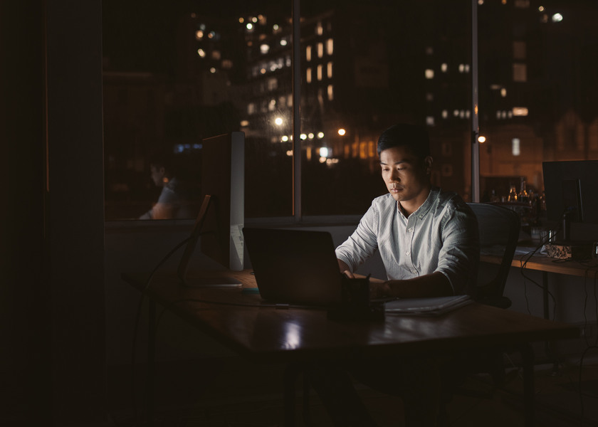 Young Asian businessman sitting at his desk working on a laptop in a dark office at night with city lights in the background