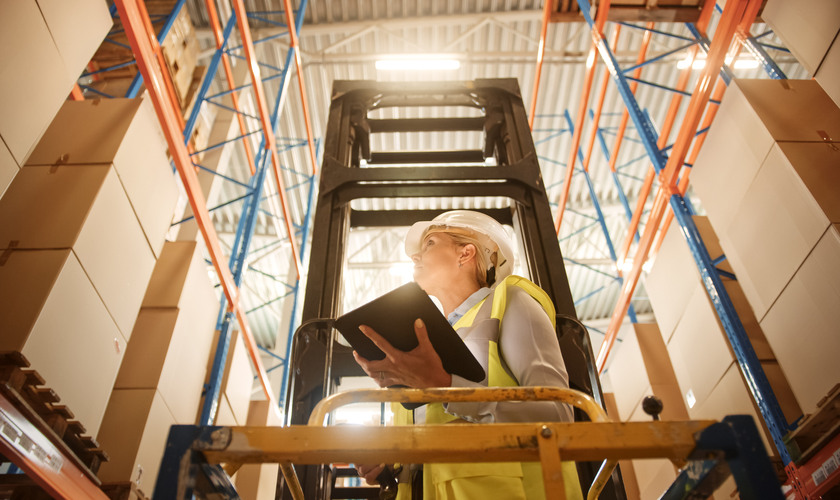 Professional Female Worker Wearing Hard Hat Lifts Herself on Aerial Work Platform to Check Stock and Inventory with Digital Tablet on the Higher Level of Retail Warehouse full of Shelves with Goods