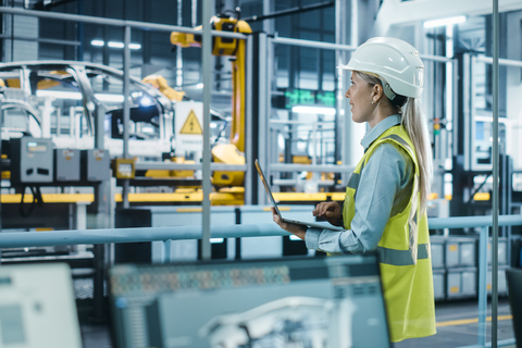 Car Factory: Female Automotive Engineer Wearing Hard Hat, Standing, Using Laptop. Monitoring, Control, Equipment Production. Automated Robot Arm Assembly Line Manufacturing Electric Vehicles.