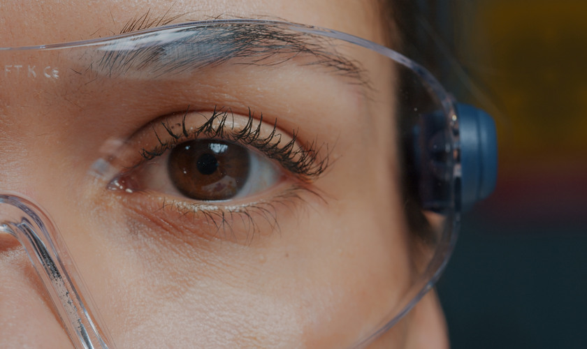 Woman with protective glasses showing one eye in front of camera, brown eye blinking and focusing sight. Person wearing safety goggles, with half of face. Genetic anatomy. Close up